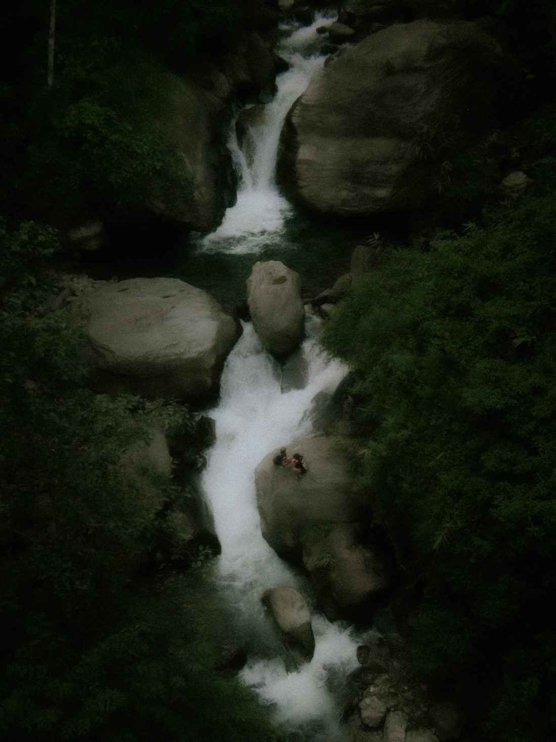 A waterfall falling between mossed boulders in dense forest, with two small figures on a rock beside it