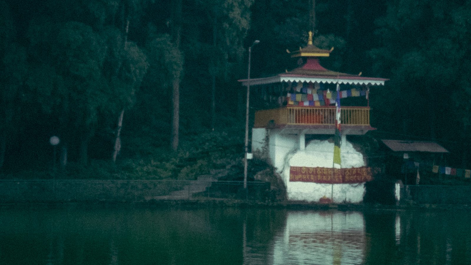 A small lakeside shrine hung with prayer flags, backed by dense forest